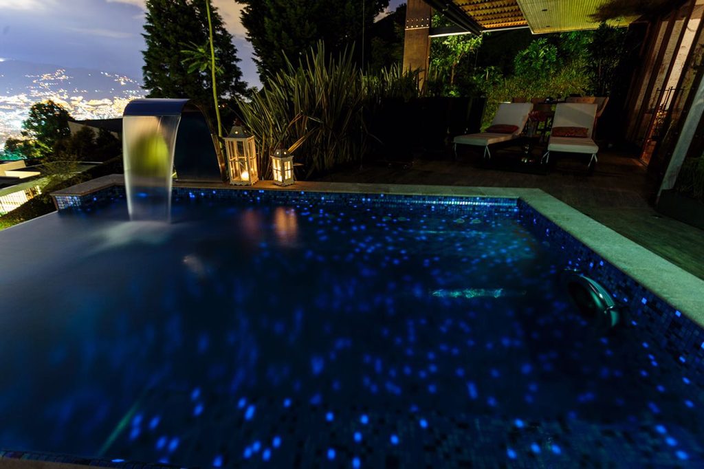 Piscina iluminada en azul al anochecer, con cascada y las luces de la ciudad al fondo